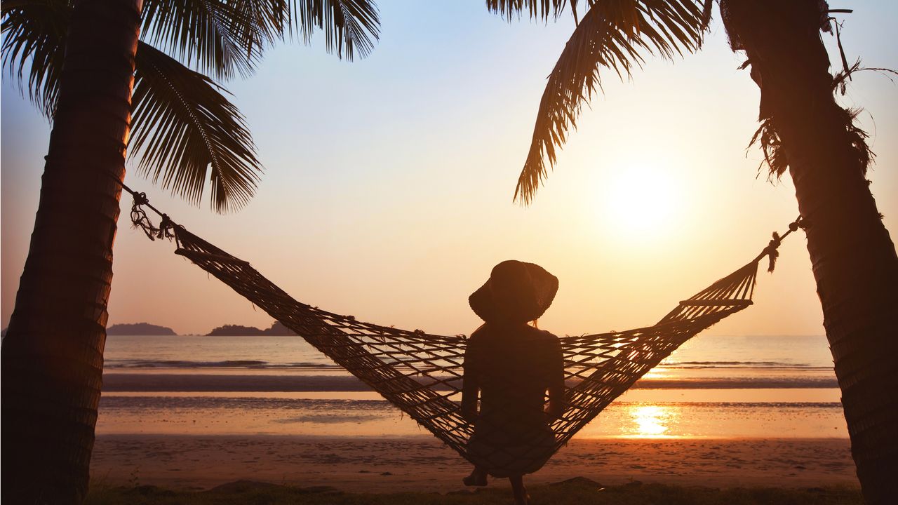 Silhouette of a woman sitting on a hammock tied between two palm trees at the beach.