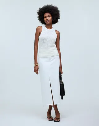 model wears white open-knit tank top and white denim maxi skirt while holding a black handbag and wearing black flip flop sandals