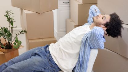 A young man relaxes in a chair, surrounded by moving boxes.