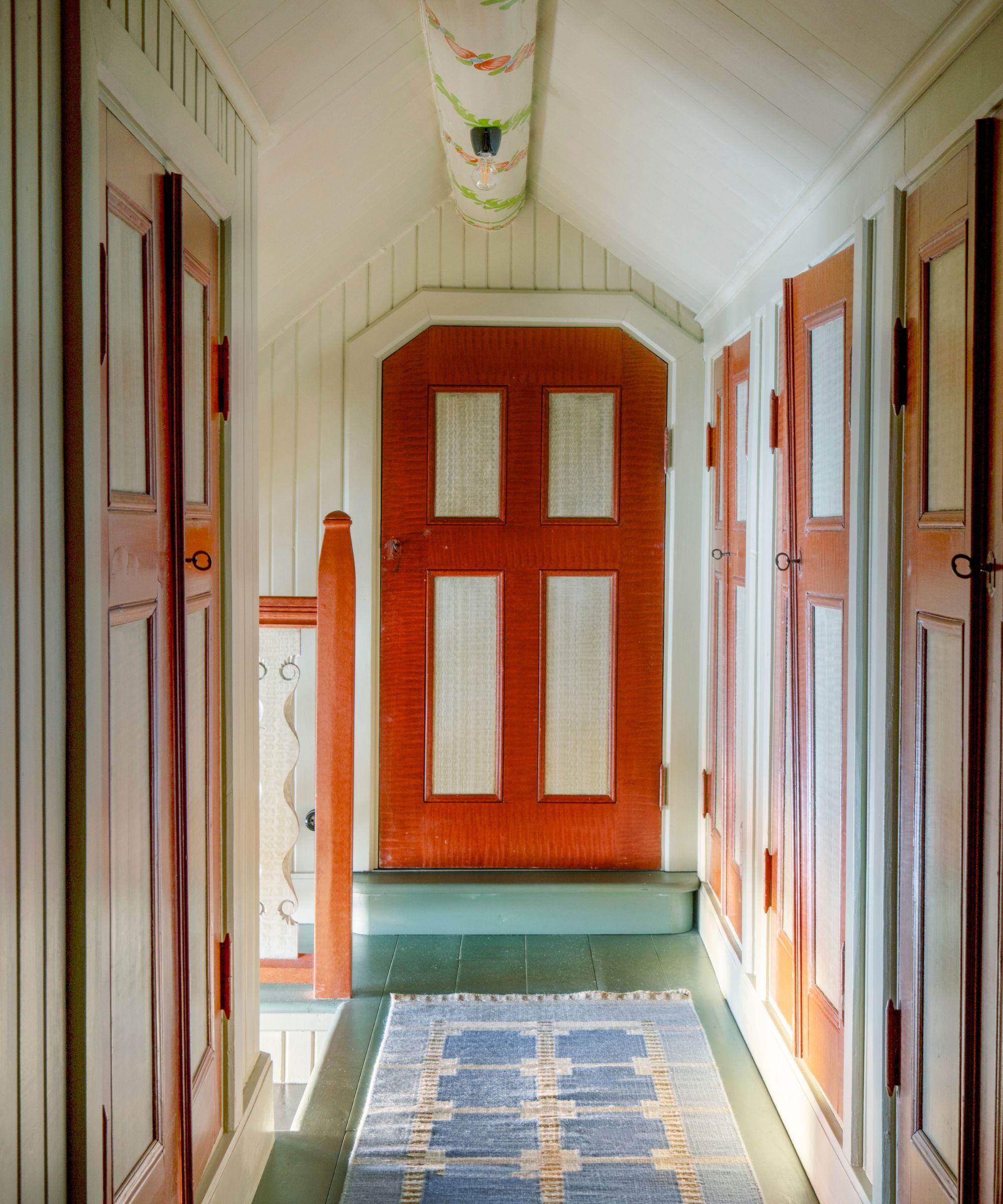 small hallway of a home with sloped ceilings with paneling and hand-painted floral motifs on the ceiling with red painted doors and a flatweave patterned rug