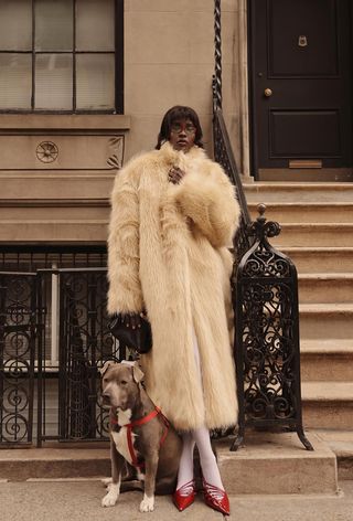 The winter capsule wardrobe item, a fur coat, is shown in a photo of a woman standing by a New York stoop with her dog wearing a long white fur coat with white tights and red pumps