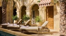 A sun-lit pool patio with rattan and beige fabric recliners, red and white striped hemming, arched passageways, and palms.
