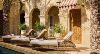 A sun-lit pool patio with rattan and beige fabric recliners, red and white striped hemming, arched passageways, and palms.