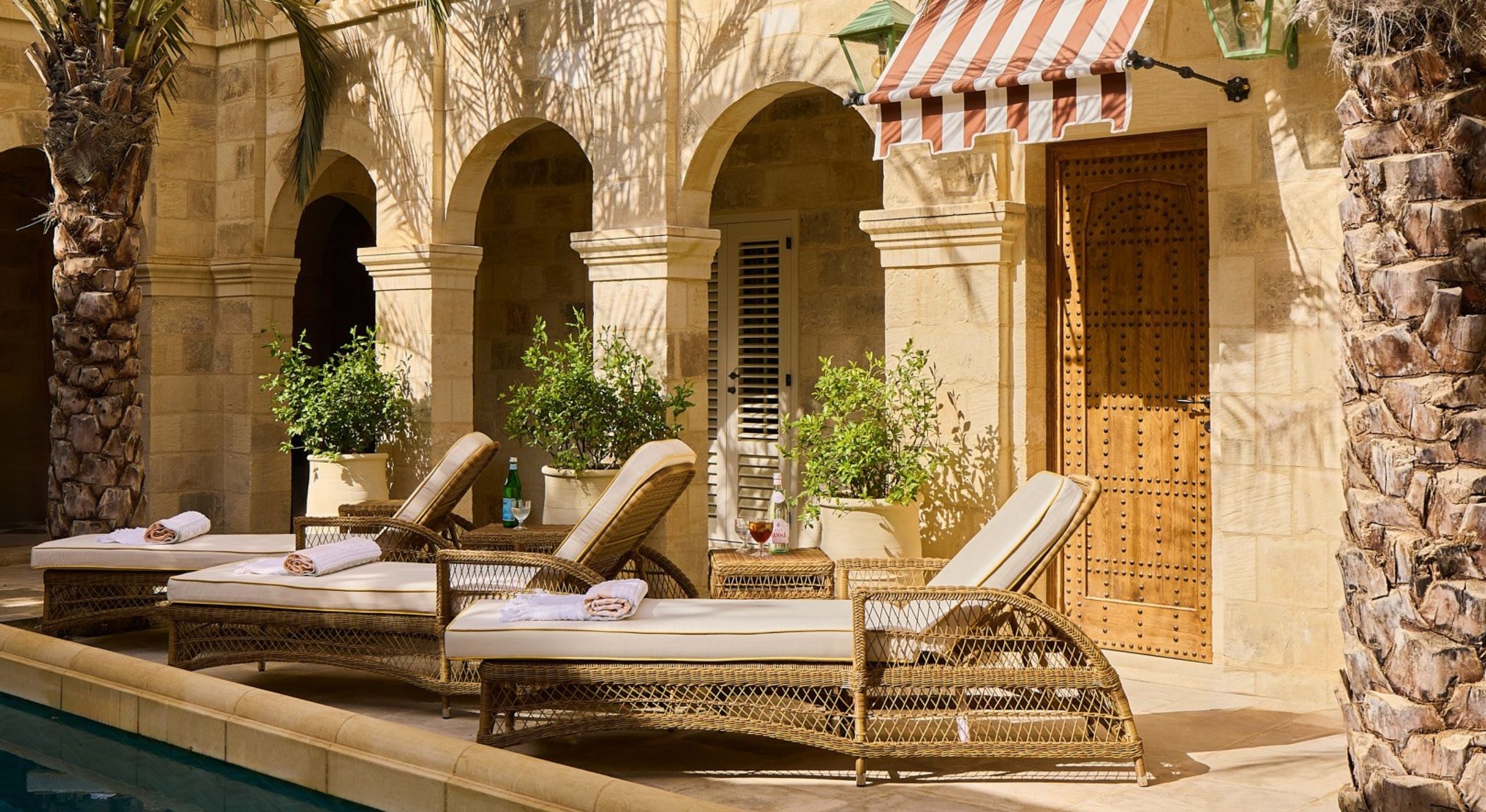 A sun-lit pool patio with rattan and beige fabric recliners, red and white striped hemming, arched passageways, and palms.