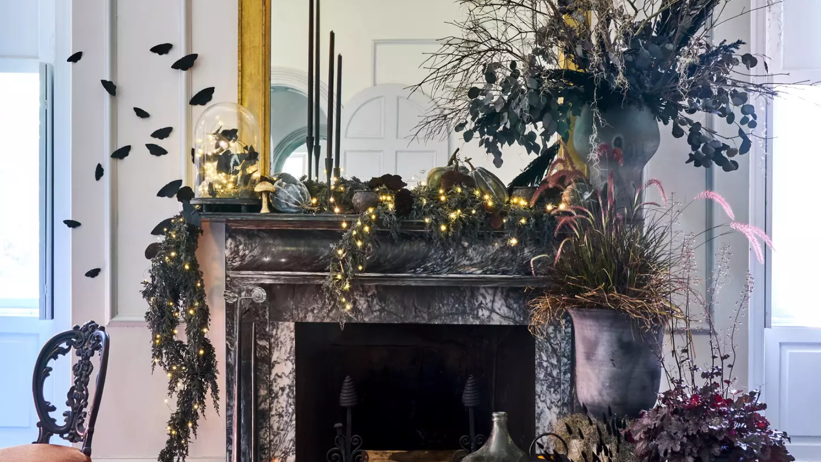 a fireplace that&#039;s been elaborately decorated for Halloween with garland, potted plants, wall-mounted moths, and decor in dark colors