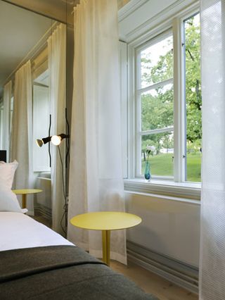 Hotel Skeppsholmen, Stockholm. A hotel room with a white bed, a round wooden coffee table and a floor lamp next to a large window with white curtains.