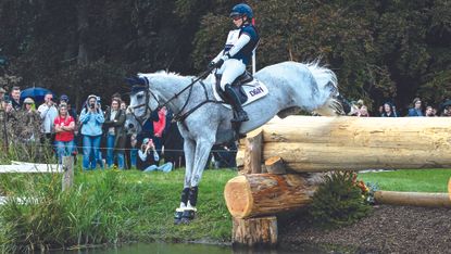 Laura Collett, a rider in a blue hat and top, rides grey horse Hester over a big log drop into water at Blenheim Horse Trials