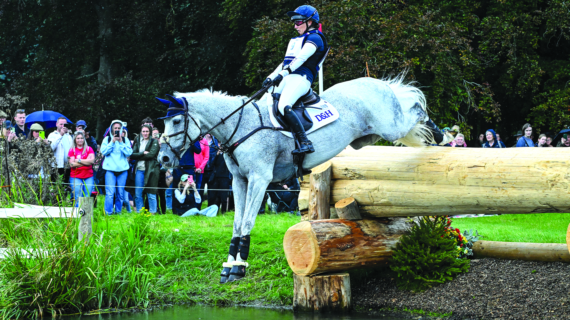 Laura Collett, a rider in a blue hat and top, rides grey horse Hester over a big log drop into water at Blenheim Horse Trials
