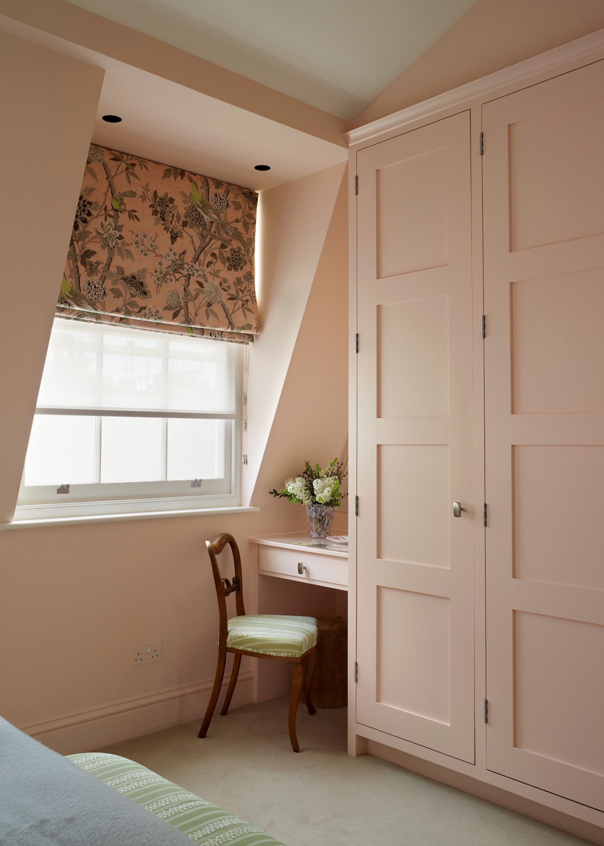 A bedroom with light pink color-drenched walls and built-in wardrobes. A window with a pink patterned roman blind, and a small vanity with a chair.