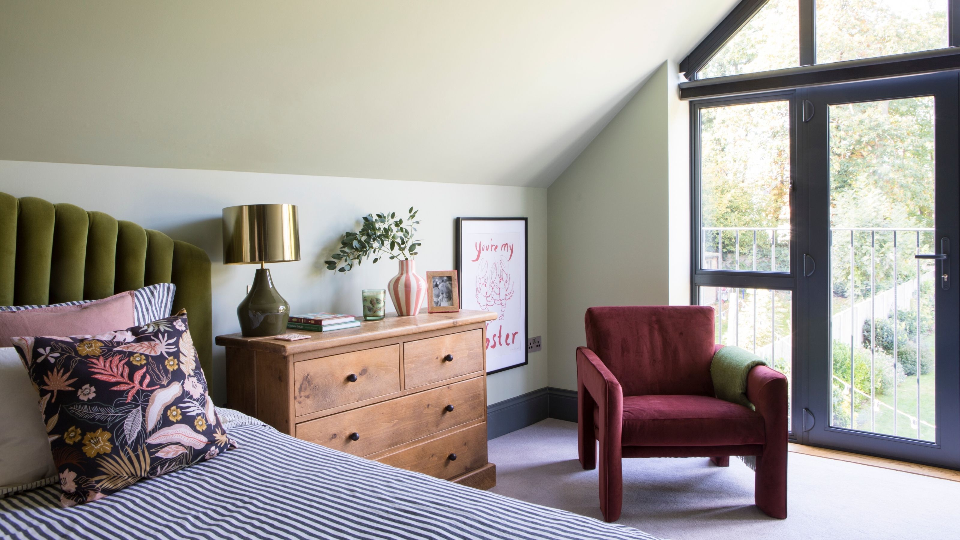 A light green bedroom with a wooden set of drawers and a burgundy armchair next to an olive green bed frame. 