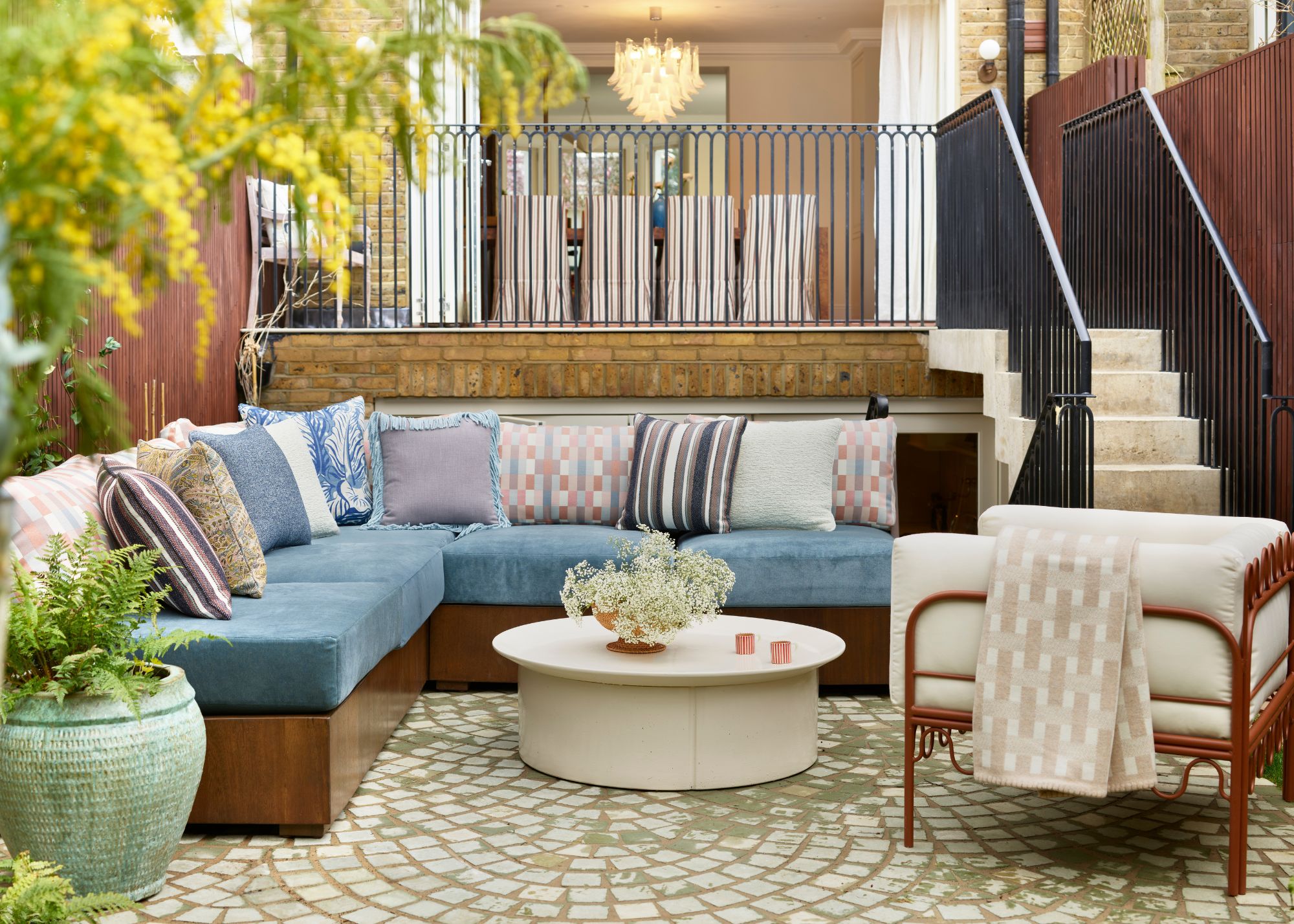 An outdoor living space with a cornflower blue corner sofa, lilac patterned cushions, a cream round coffee table, and a modern minimalist armchair with a rust-colored frame and cream cushion.