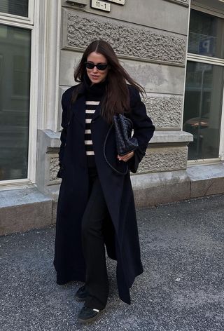 A black turtleneck outfit is shown in a photo of a French woman standing on the sidewalk wearing a navy trench coat over a black striped turtleneck sweater with black trousers, black samba sneakers, and a black Bottega Veneta bag