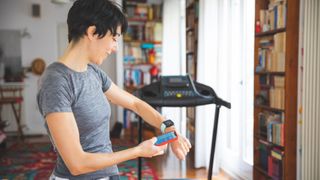 Woman looking at fitness tracker and phone, standing at the end of a treadmill at home