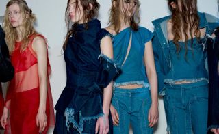 Row of four young female models, one in red mesh clothing, three in slashed and cut off denim outfits, heavy eye makeup, messy hair styles, white walls