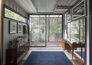 Inside a small room with shelf units and a side board. Looking out to an enclosed balcony covered by perforated panels with greenery in the distance.