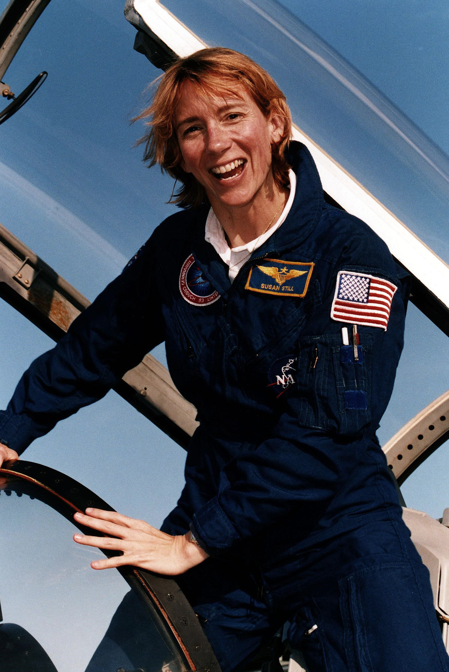 woman is standing next to the open canopy of an airplane cockpit.  she is wearing a flight suit