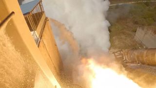 flames shoot out of a cone-shaped rocket engine poking out of a hangar-like building