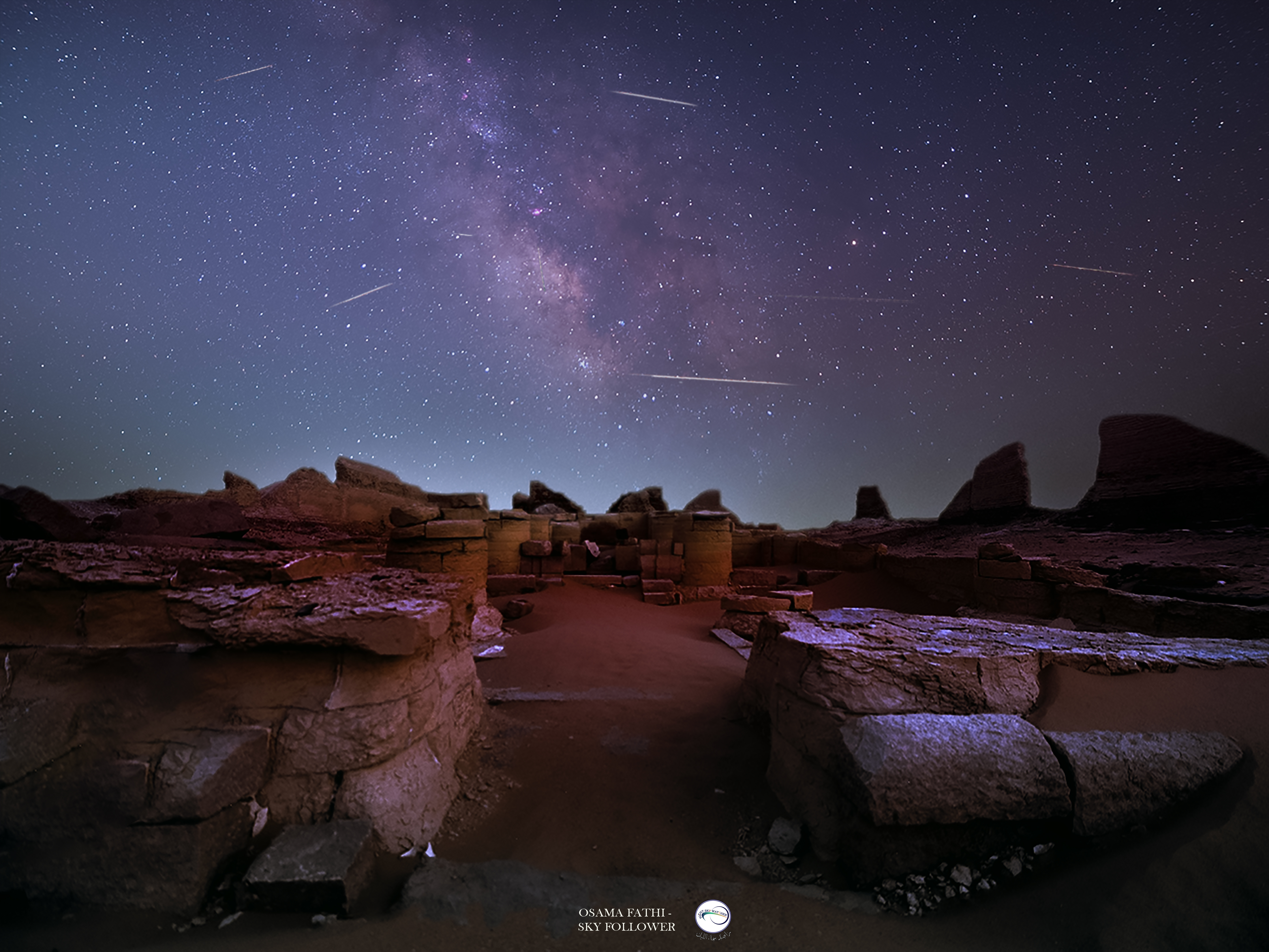 Shooting stars are pictured as lines streaking across the glowing band of the Milky Way, which is visible tumbling vertically towards the horizon. Stone ruins are visible below and a soft glow is detectable near the horizon.