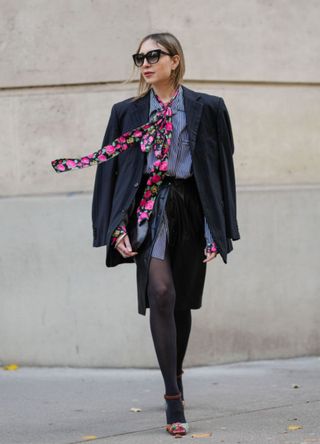 Emy Venturini wears sunglasses, a striped shirt from Balenciaga, a black oversize blazer jacket, a pink colored floral print tie, a knee length black leather skirt from Patrizia Pepe, black tights from Wolford, a black leather clutch bag from Pierre Cardin, sunglasses from Dita, colored embellished rhinestones shoes from Rene Caovilla, earrings from Emy x Cloel, during a street style fashion photo session, on November 20, 2024 in Paris, France