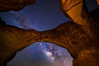 A large rock arch is in the foreground and in the background is the milky way.