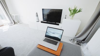 Macbook open on grey sofa opposite small TV and soundbar on white unit