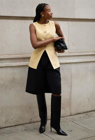 A woman wearing gold earrings, a yellow waistcoat, a black clutch bag, black Bermuda shorts, and black knee-high boots.