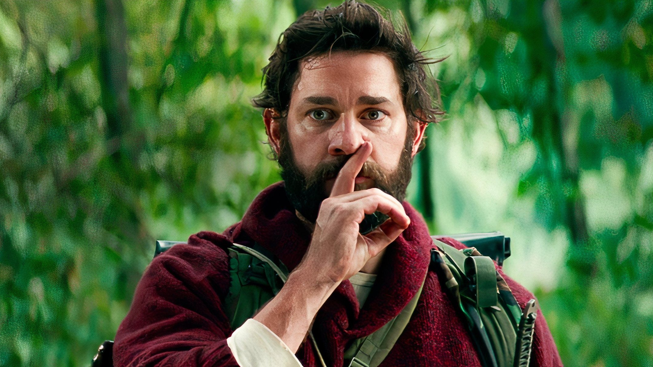 A bearded man with a backpack holds his hand over his mouth making a shush motion