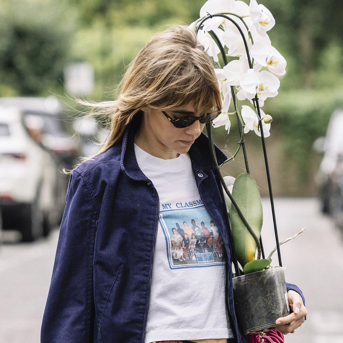 Suki Waterhouse wearing a navy jacket, white T-shirt, and burgundy shorts and sneakers while carrying a plant in London