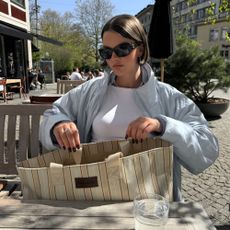 A woman sat outside in the sunshine with a jacket and sunglasses on