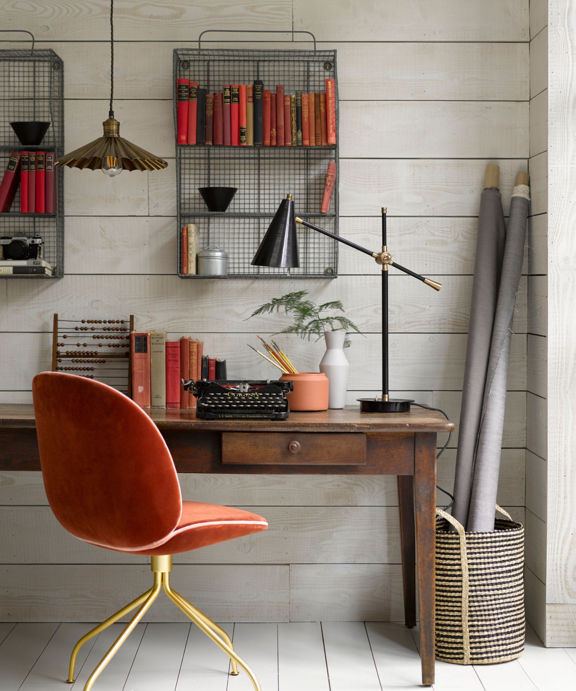 Eclectic home office with a vintage wooden desk, burnt orange velvet chair, black desk lamp, and wire wall shelves filled with red and orange books.