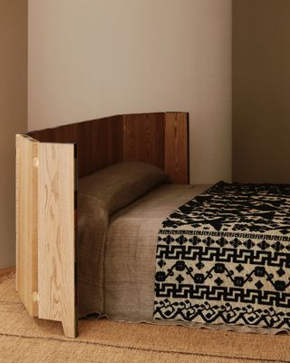 A bedframe shaped out of foldable wood adds a touch of rusticness to an earthy-shaped bedroom featuring brown and black linen bedding and a rattan carpet.