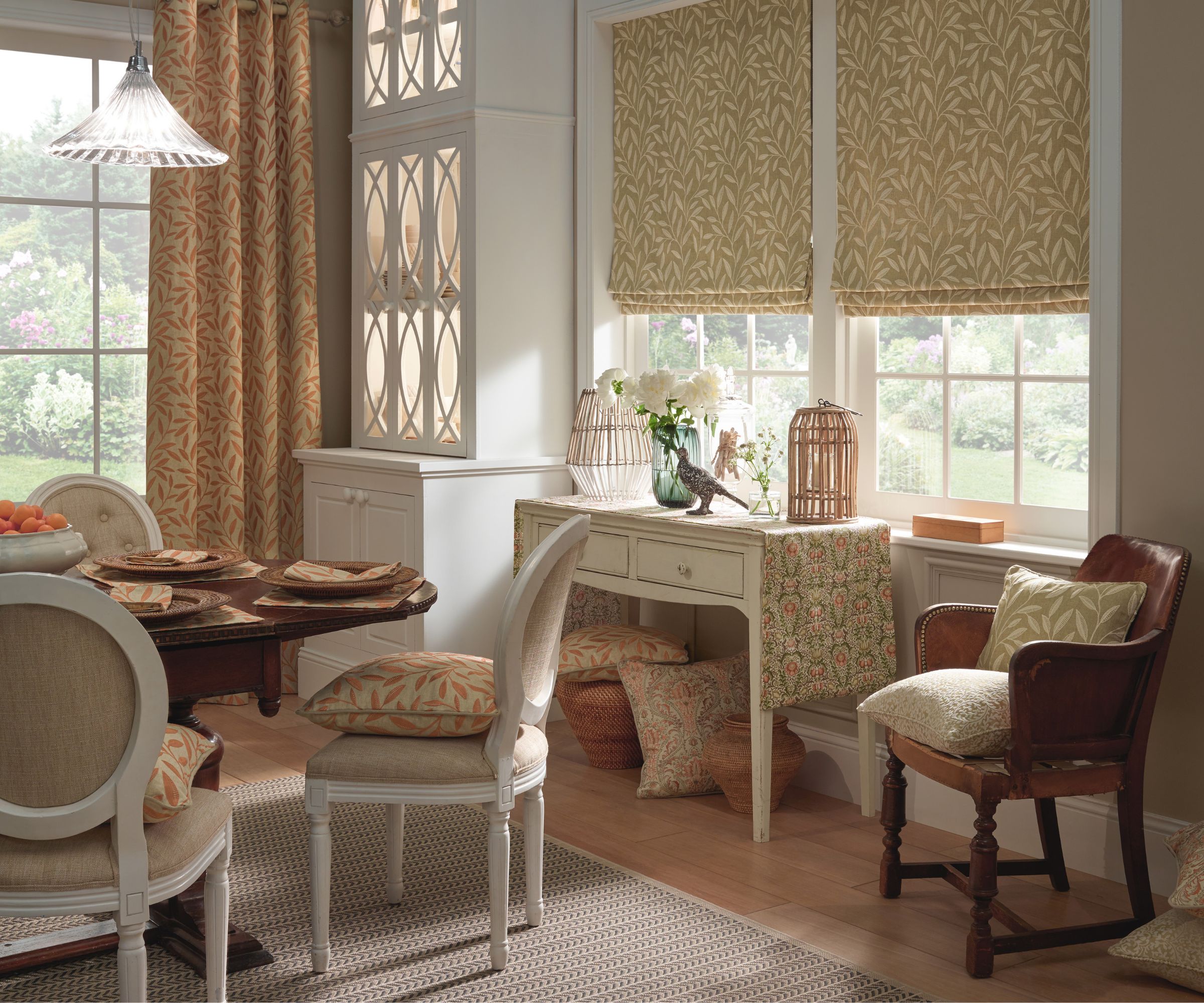 A traditional dining room with patterned Roman blinds, matching curtains and soft neutral tones. A small desk sits beneath the window, surrounded by cushioned chairs, woven baskets and floral accessories.
