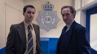 Sam Swainsbury as DI Clive Roscoe and Bertie Carvel as Dalgliesh stand in front of an East Essex Police sign in Dalgliesh.