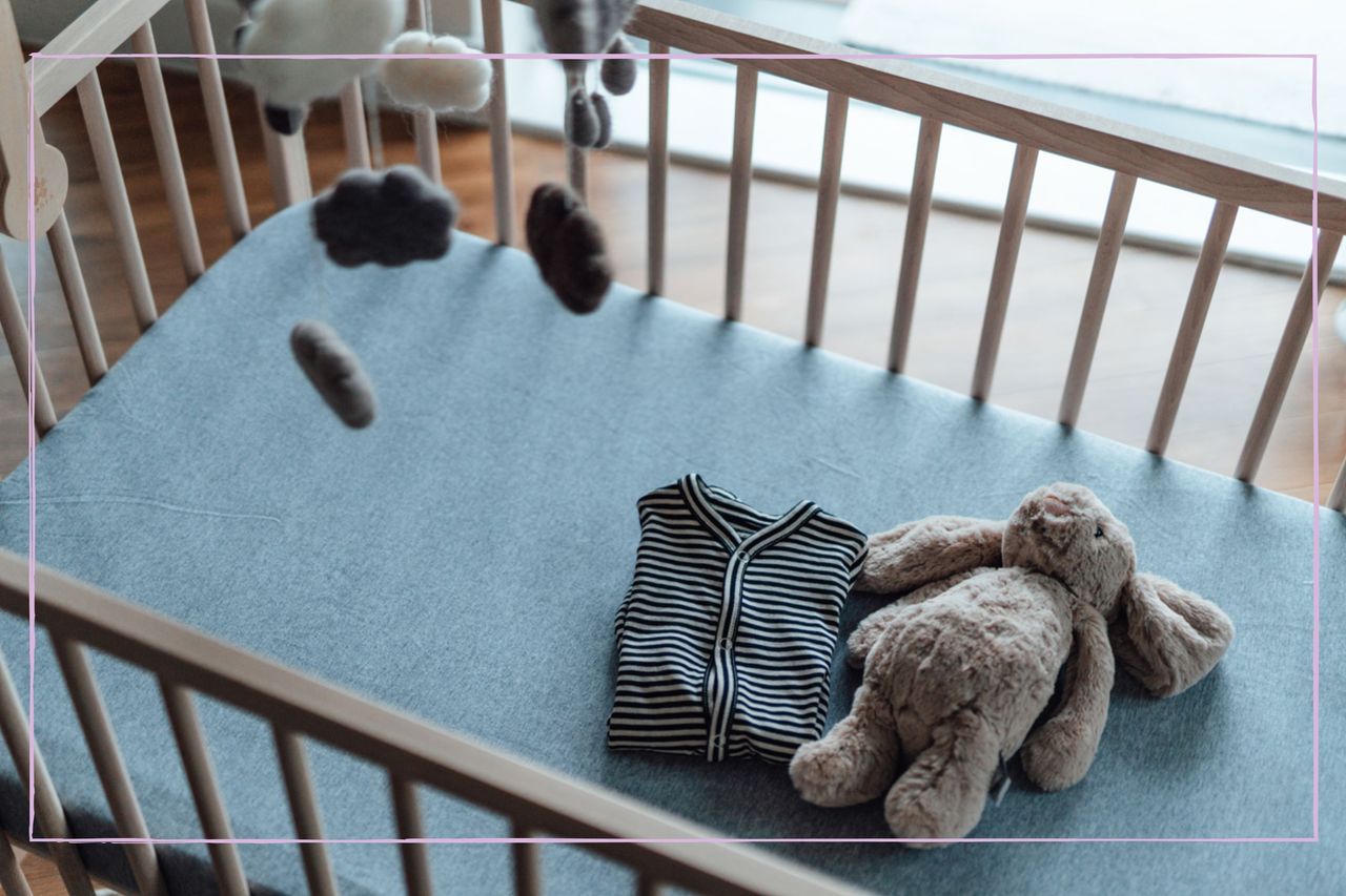 An empty cot with a folded baby grown and toy rabbit inside