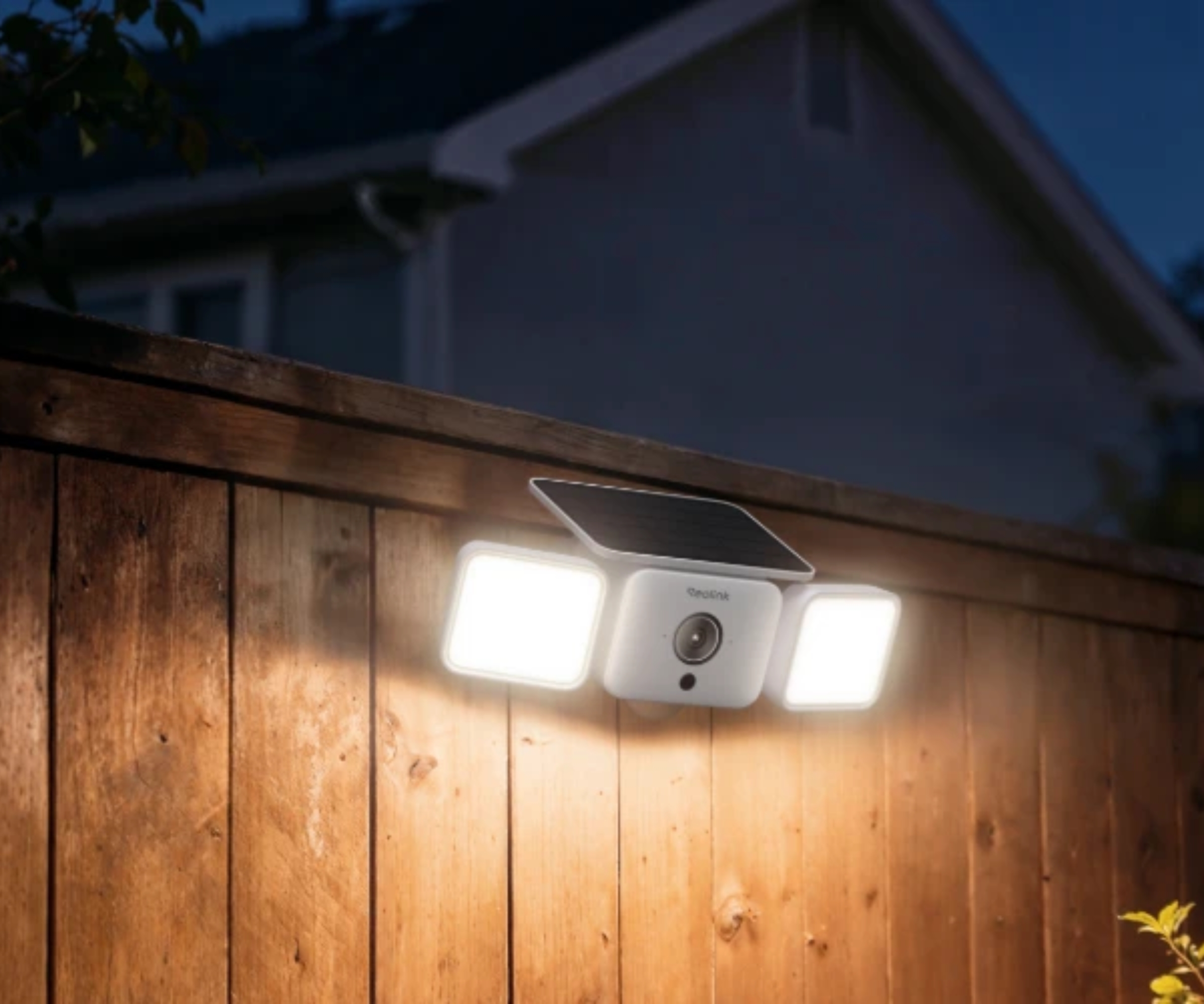 Reolink solar floodlight camera lit up on wooden fence at dusk, house silhouetted in background.