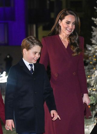 Prince George wearing a suit and tie smiling walking next to mom Kate Middleton past a Christmas tree at the 2022 Together at Christmas concert