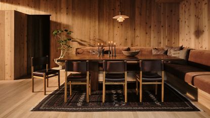Image of a dark rug on a light wooden floor. The rug is under a dark dining table with a corner bench seat upholstered in burnt orange velvet fabric and black leather chairs on the other side.