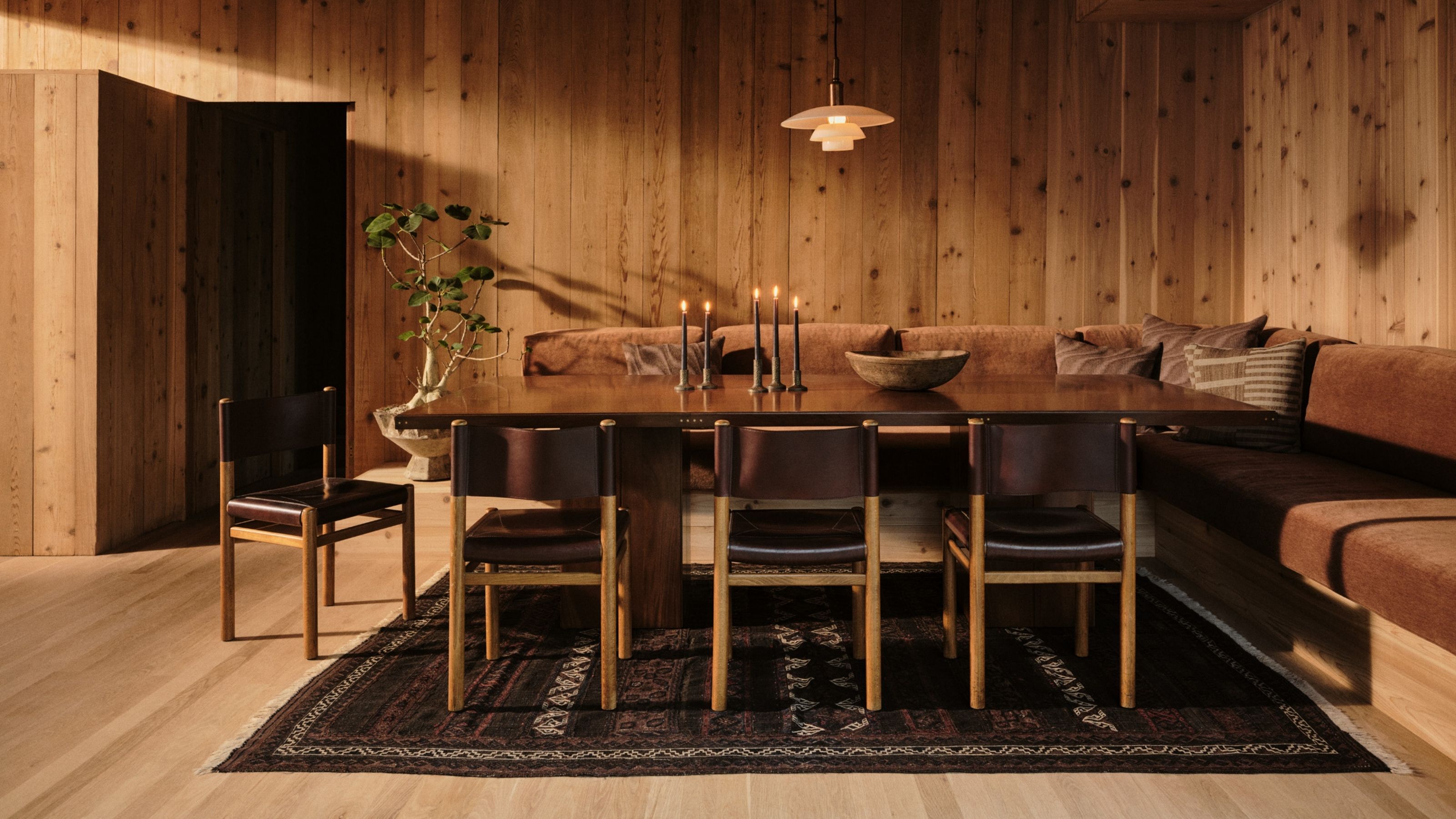 Image of a dark rug on a light wooden floor. The rug is under a dark dining table with a corner bench seat upholstered in burnt orange velvet fabric and black leather chairs on the other side.