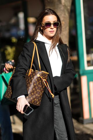 Woman wears Louis Vuitton Neverful Bag with white hoodie, black jeans, and black trench coat.