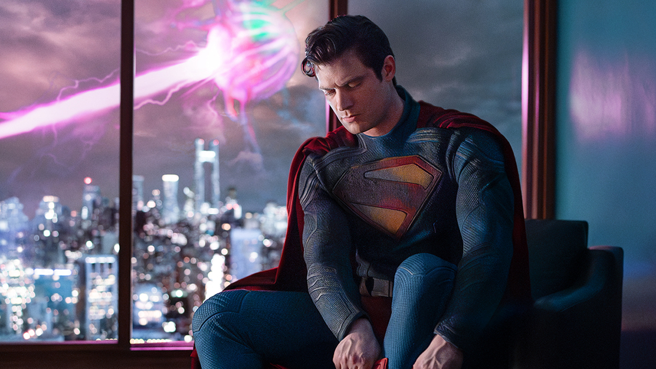 Captura de pantalla de Man of Steel de David Corenswet vistiendo un traje en un apartamento en Metrópolis mientras un láser rosa dispara de fondo en la película Superman de 2025.