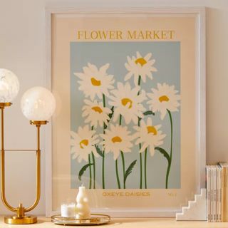 A Gale Switzer Flower Market Oxeye Daisies Art Print on top of a cabinet next to a lamp, books, and candles