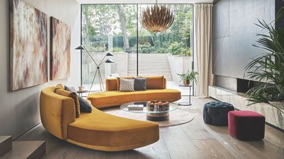A bright living room seating area with wooden flooring, two matching curved yellow sofas, abstract artworks on the wall, two velvet pouffes and large glass windows showing a leafy garden with stone stairs behind.