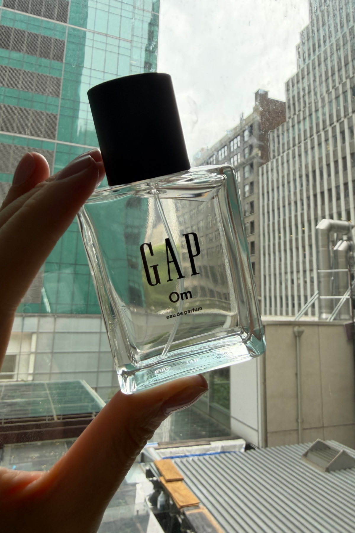 a hand holding up a clear glass bottle of Gap Om perfume to the window