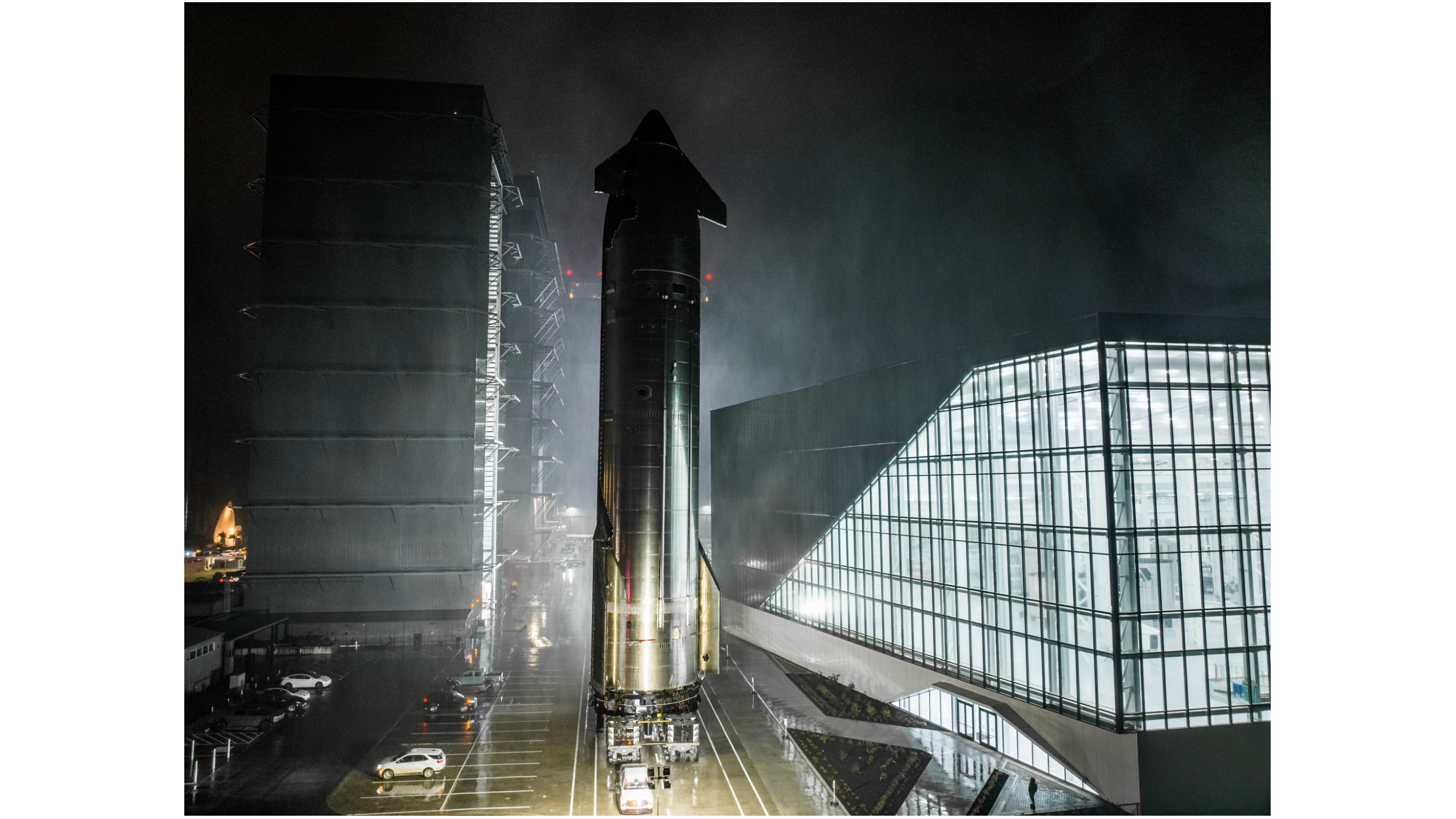 A large silver rocket rolls down a road at night,passing close to a lit-up building