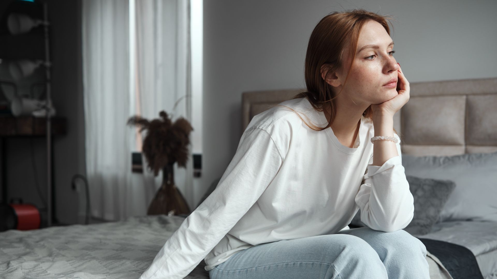 A woman sits on the edge of her bed looking sad.