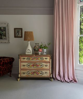 Traditional bedroom with gray carpet, painted wooden chest of drawers, decorated with table lamp, pink curtains, large floor to ceiling window, white painted walls, gray painted feature