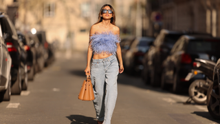 Angela Gonzalez wears black sunglasses, silver earrings, a purple feather shoulder-off /cropped top, blue faded denim mom jeans pants, a brown camel shiny leather handbag, dark brown shiny leather strappy heels sandals / shoes, during a street style fashion photo session, on March 26, 2022 in Paris, France.