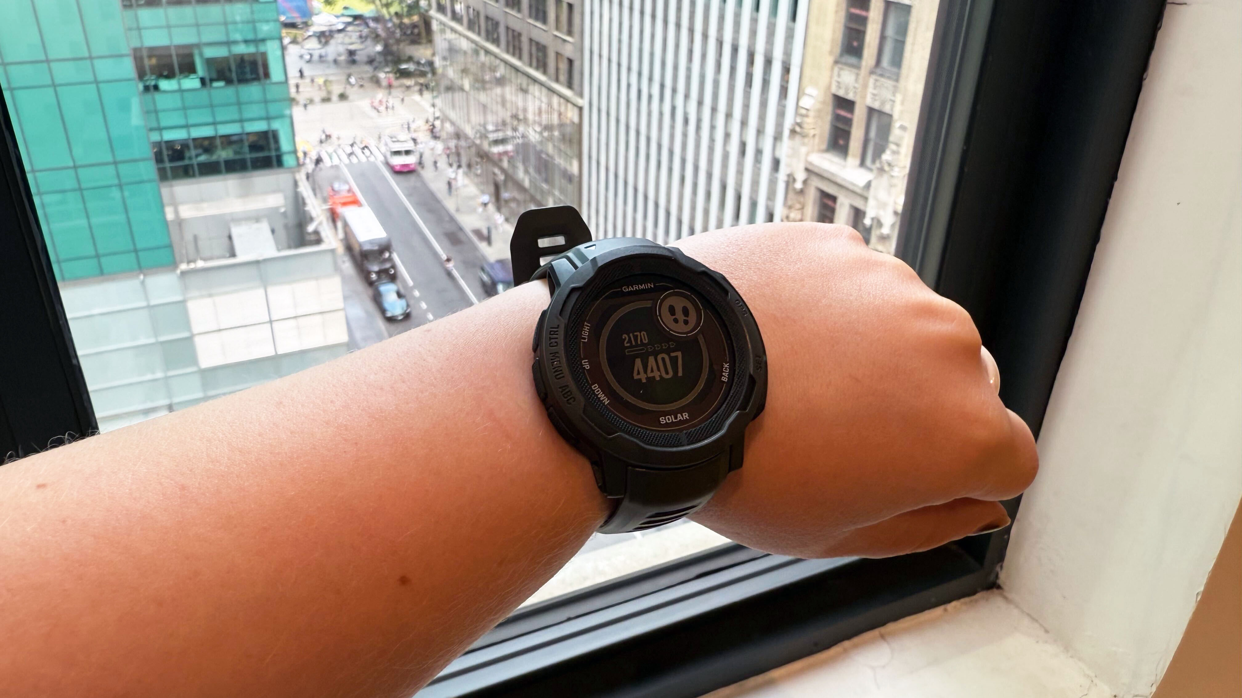 Garmin Instinct 2 Solar on left wrist, in front of a window overlooking Bryant Park in New York City.