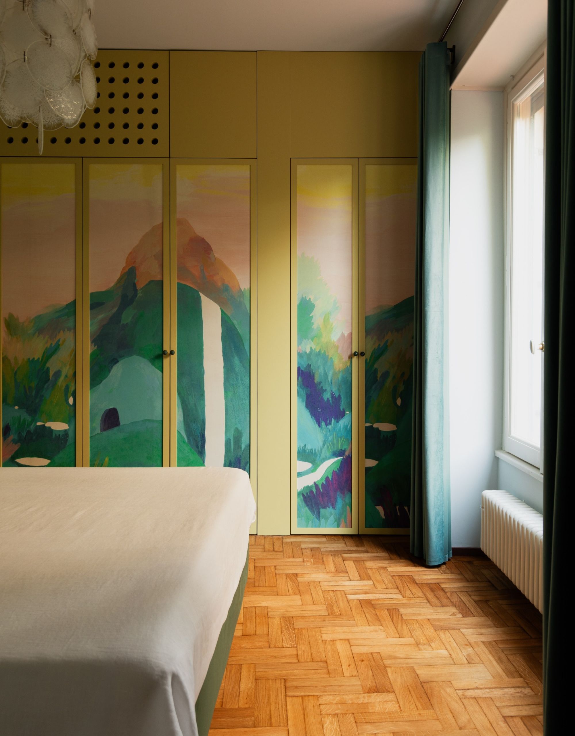 A bedroom with parquet flooring, a simple bed and a wardrobe painted in a bright mural