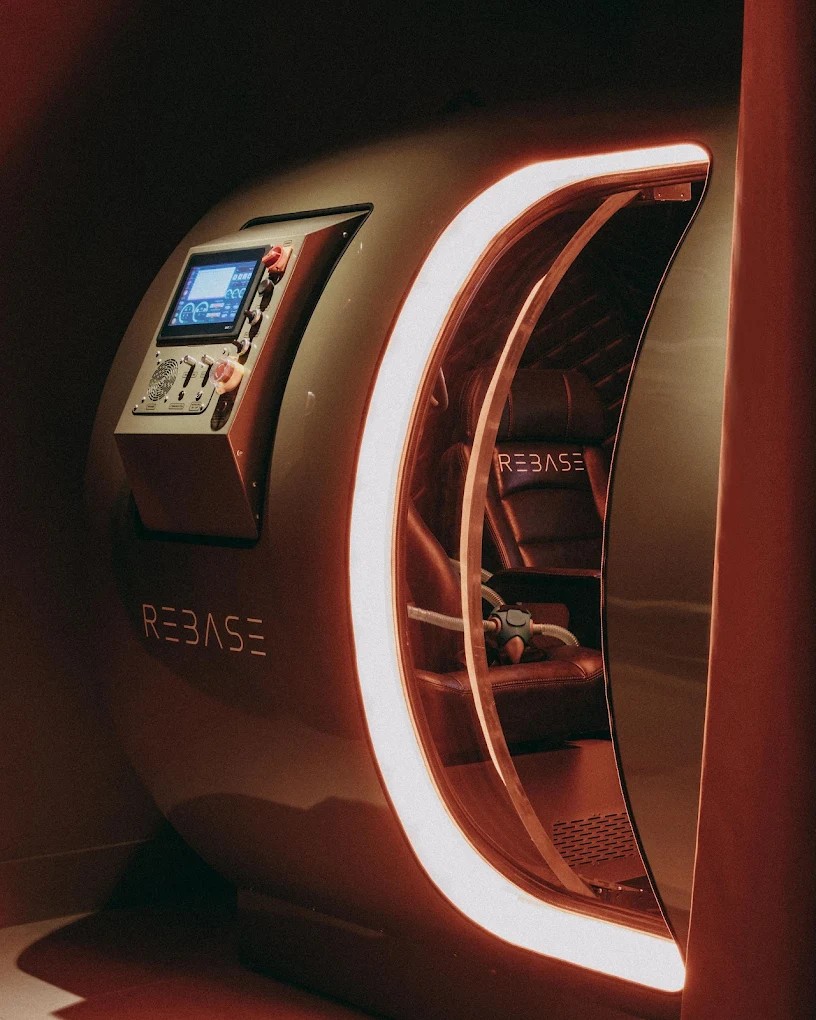 A warmly decorated, contemporary sauna hub with creamy and terracotta plaster walls, curvilinear upholstered furniture with green fabric, wooden details, and warm pendant as well as spherical sconce lighting throughout.